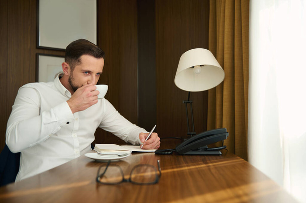 Portret przystojnego, atrakcyjnego biznesmena w średnim wieku, przedsiębiorcy, dziennikarza, pisarza trzymającego srebrny długopis i notującego w notatniku pijąc pyszną świeżo parzoną kawę w pokoju hotelowym - Zdjęcie, obraz