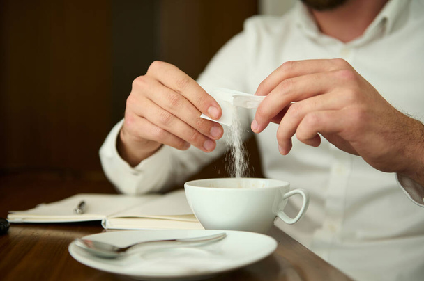 Großaufnahme eines bis zur Unkenntlichkeit verkleideten Mannes in weißem Hemd, der Zucker aus Stöcken in eine weiße Keramiktasse mit einem frisch gebrühten Kaffeegetränk streut, an einem Holztisch sitzt und eine Kaffeepause genießt. Ernte - Foto, Bild