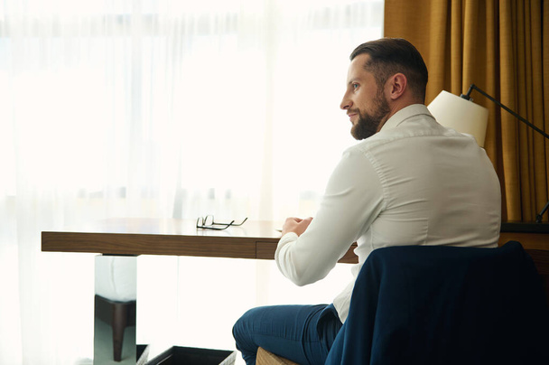 Οπίσθια όψη μεσήλικα επιχειρηματία που κάθεται σε ένα τραπέζι σε δωμάτιο ξενοδοχείου - Φωτογραφία, εικόνα