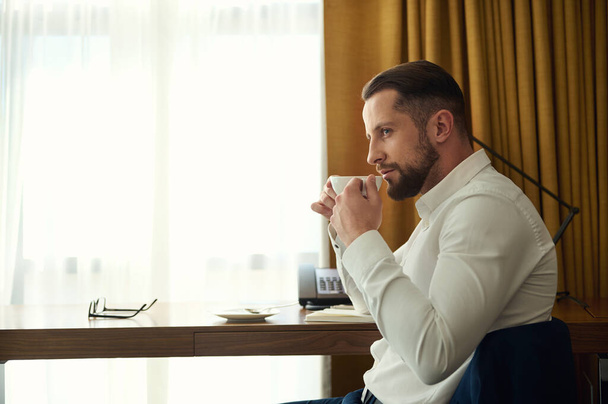 Handsome self-confident European businessman, entrepreneur on business trip drinking coffee, sitting at a desk in hotel room - Фото, изображение