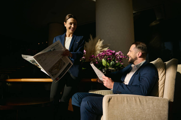Two handsome people, middle aged Caucasian man and beautiful elegant woman in business suit, colleagues, business partners on business trip, talking and reading a newspaper in the hotel lounge zone - Photo, Image