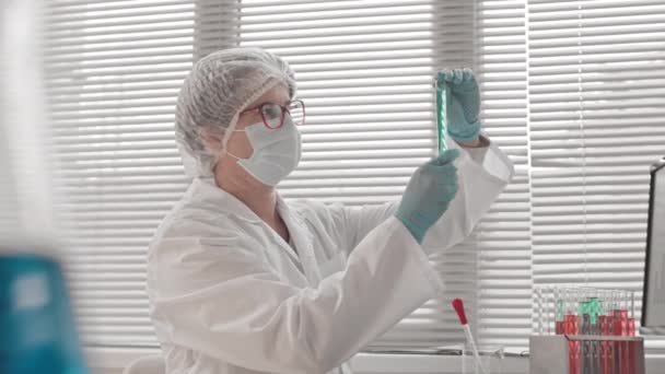 Niski kąt widzenia z boku inteligentnej białej kobiety noszącej medyczną suknię i maskę, siedzącej za biurkiem w centrum badawczym w dzień, pracującej z chemikaliami w probówkach - Materiał filmowy, wideo
