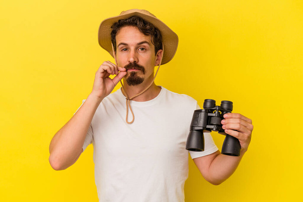 Young caucasian man holding binoculars isolated on yellow background with fingers on lips keeping a secret. - Photo, Image