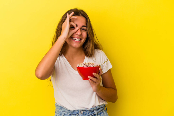 Joven mujer caucásica sosteniendo cereales aislados sobre fondo amarillo excitado manteniendo el gesto bien en el ojo. - Foto, imagen