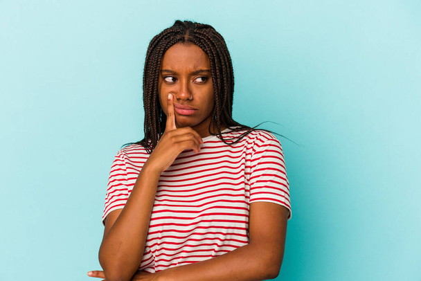 Молода афроамериканська жінка, ізольована на синьому фоні, дивиться в бік з сумнівним і скептичним виразом.. - Фото, зображення