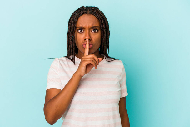 Νεαρή Αφροαμερικανή, απομονωμένη σε μπλε φόντο, κρατώντας μυστικό ή ζητώντας σιωπή.. - Φωτογραφία, εικόνα