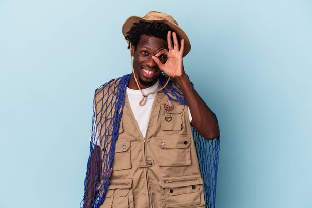 Young african american fisherman holding a net isolated on blue background excited keeping ok gesture on eye. - Φωτογραφία, εικόνα