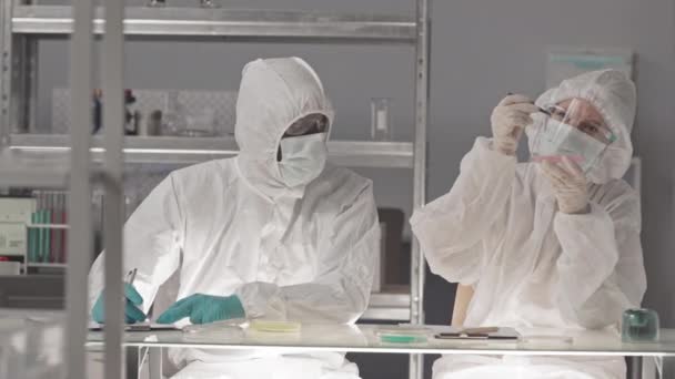 Medium long of male African American and female Caucasian laboratory workers wearing hazmat overalls, goggles and masks, sitting in research center, analyzing biological material in Petri dishes - Footage, Video
