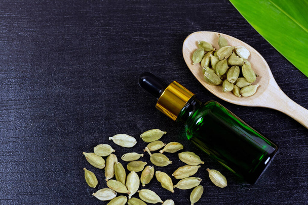 Cardamom essential oil in green glass bottle with dry cardamom spice on wooden spoon and green leaf on black wooden background, top view. - Foto, Imagem
