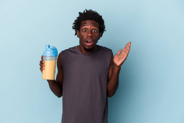 Молодой африканский американец, держащий протеиновый молочный коктейль на синем фоне удивлен и шокирован. - Фото, изображение