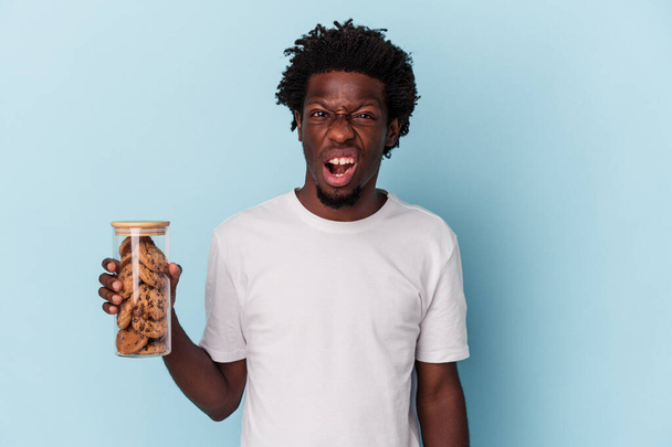 Nuori afrikkalainen amerikkalainen mies pitelee suklaakeksejä sinisellä taustalla huutaen hyvin vihainen ja aggressiivinen. - Valokuva, kuva
