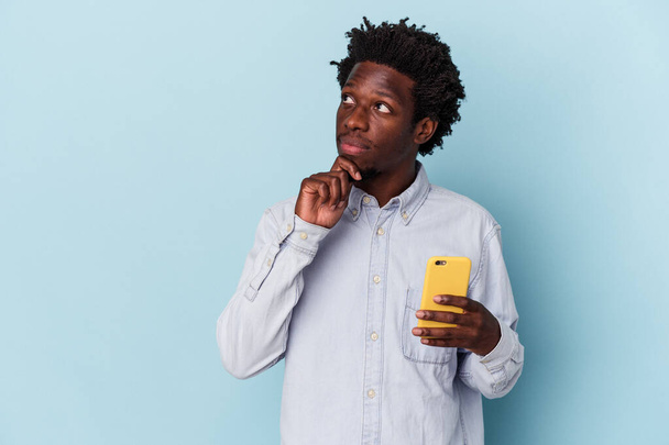 Elinde cep telefonu olan Afrikalı genç bir adam mavi arka planda izole edilmiş şüpheli ve şüpheci bir ifadeyle yana bakıyor.. - Fotoğraf, Görsel