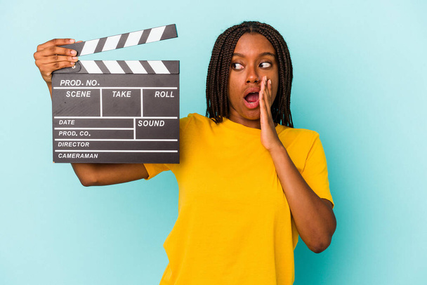 Giovane donna afroamericana che tiene un clapperboard isolato su sfondo blu sta dicendo una notizia segreta frenante caldo e guardando da parte - Foto, immagini