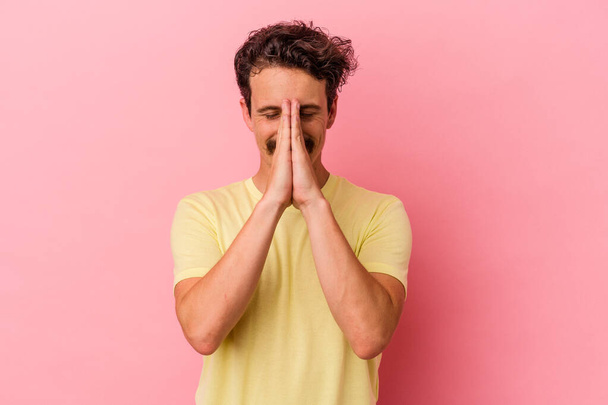 Jonge blanke man geïsoleerd op roze achtergrond hand in hand bidden in de buurt van mond, voelt vertrouwen. - Foto, afbeelding
