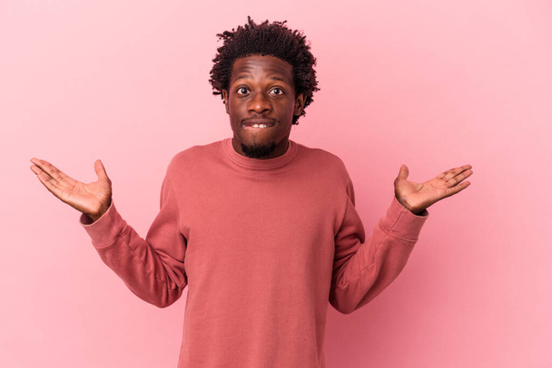 Young african american man isolated on pink background confused and doubtful shrugging shoulders to hold a copy space. - 写真・画像