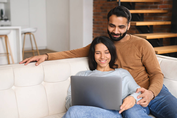 Young Pakistani couple using laptop while sitting on the sofa at home. Spouses watching movie on laptop. Happy man and woman embracing while relaxing - Foto, imagen