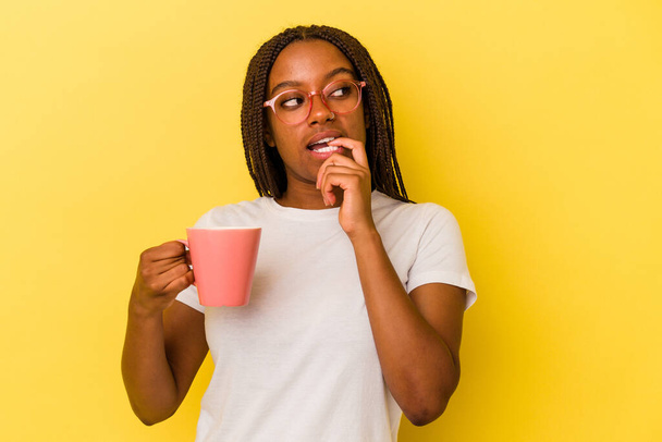 Young african american woman holding a mug isolated on yellow background  relaxed thinking about something looking at a copy space. - Foto, Imagem