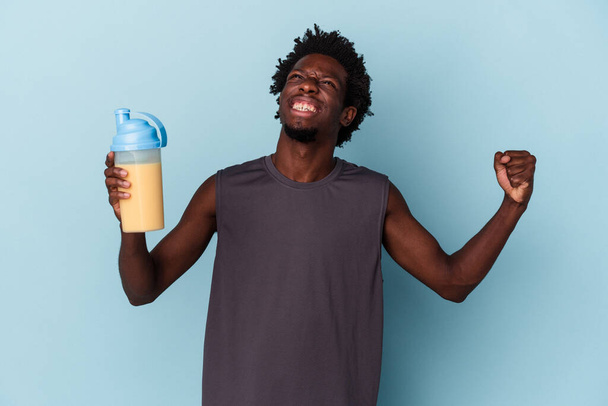 Young african american man holding protein milkshake isolated on blue background - Photo, image