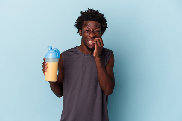 Nuori afrikkalainen amerikkalainen mies pitelee proteiinipirtelöä, joka on eristetty sinisellä taustalla puree kynsiä, hermostunut ja hyvin ahdistunut.. - Valokuva, kuva