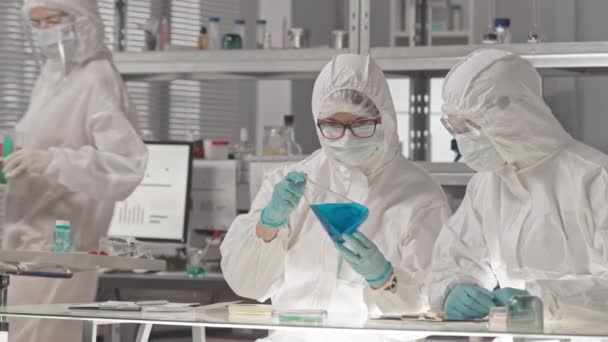 Valkoihoisten mies- ja naispuolisten laboratoriotyöntekijöiden vyötärö, jolla on yllään suojahaalarit, suojalasit ja naamarit, istuu tutkimuskeskuksessa analysoimassa vaarallisia kemikaaleja koeputkissa - Materiaali, video
