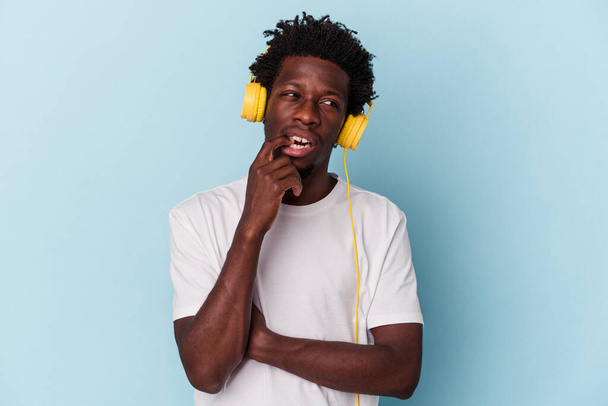 Hombre afroamericano joven escuchando música aislado sobre fondo azul relajado pensando en algo mirando un espacio de copia. - Foto, imagen