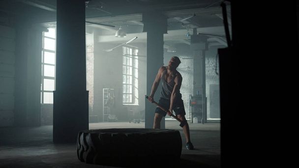 Guy doing strength exercise in sport club. Man beating big tire with hammer - Zdjęcie, obraz