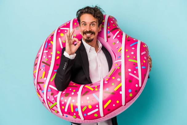 Jeune homme d'affaires avec donut gonflable isolé sur fond bleu joyeux et confiant montrant un geste correct. - Photo, image