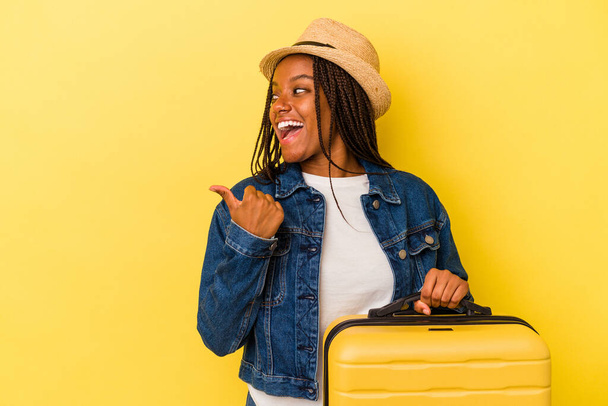 Young african american traveler woman holding suitcase isolated on yellow background  points with thumb finger away, laughing and carefree. - 写真・画像