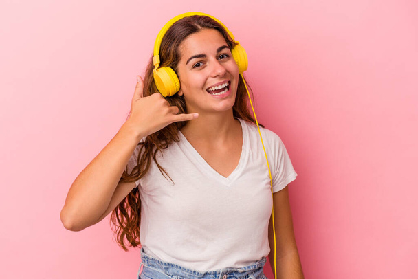 Jonge blanke vrouw luisteren naar muziek geïsoleerd op roze achtergrond tonen een mobiele telefoon oproep gebaar met vingers. - Foto, afbeelding