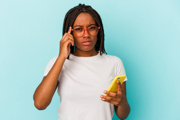 Молодая африканская американка держит мобильный телефон изолированный на синем фоне указывая висок пальцем, думая, сосредоточена на задаче. - Фото, изображение