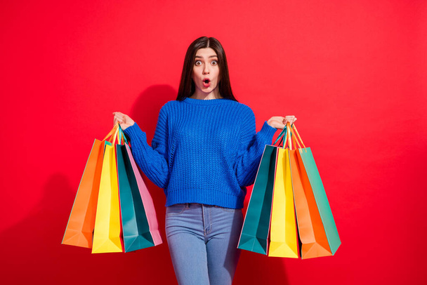 Photo of amazed shocked surprised young woman hold hands bags buy clothes isolated on red color background - Foto, Imagen
