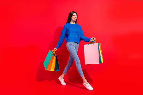 Full size profile side photo of attractive happy young woman look empty space hold bags shop isolated on red color background - Foto, afbeelding