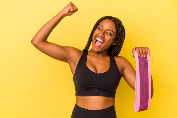 Jeune femme sportive afro-américaine tenant une bande élastique isolée sur fond jaune levant le poing après une victoire, concept gagnant. - Photo, image