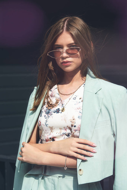 Güneş gözlüğü takan genç bir kadının moda portresi, üst ve mavi takım elbise. Genç, güzel, mutlu model, karanlık arka planda poz veriyor. - Fotoğraf, Görsel