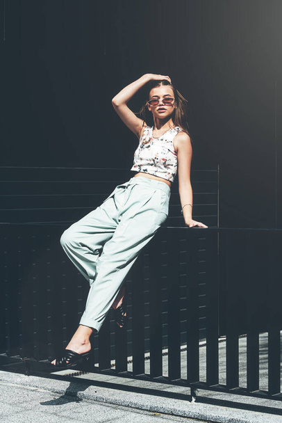 Retrato de moda de mujer joven con gafas de sol, top, slingbacks, traje azul. Joven modelo feliz hermosa posando cerca de rejilla de metal gris - Foto, Imagen