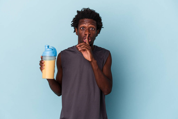 Молодой африканский американец, держащий протеиновый коктейль, изолированный на синем фоне, хранит секрет или просит тишины. - Фото, изображение