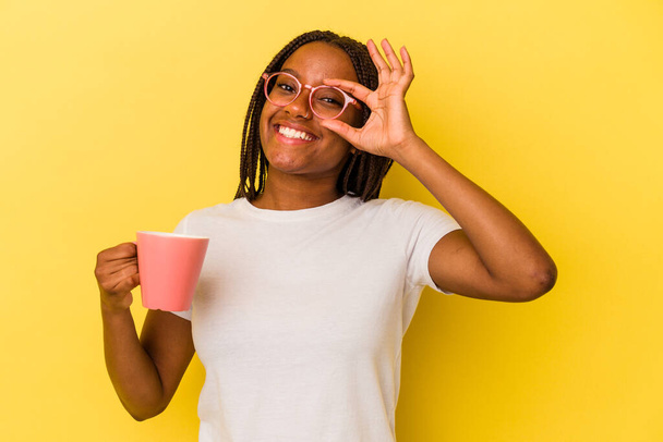 Joven afroamericana americana sosteniendo una taza aislada sobre fondo amarillo excitada manteniendo el gesto de bien en el ojo. - Foto, Imagen