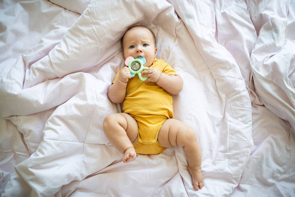 Caucasian blonde baby seven months old lying on bed at home. Kid wearing cute clothing trendy colors: illuminating yellow. Infant playing with toys - Photo, image