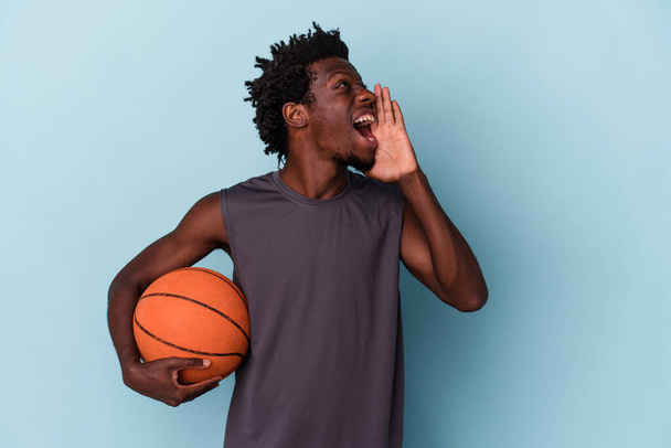 Young african american man playing basketball isolated on blue background shouting and holding palm near opened mouth. - Photo, image