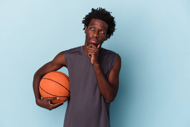 Young african american man playing basketball isolated on blue background looking sideways with doubtful and skeptical expression. - 写真・画像