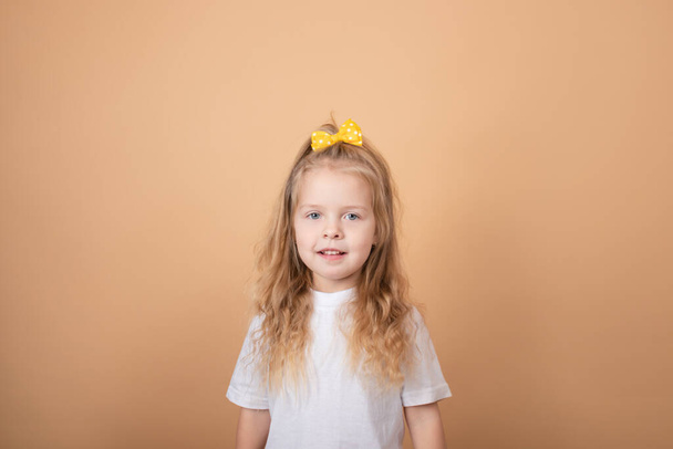 portrait of a sweet blonde little girl in white t-shirt and with yellow bow on her head. on brown background - Foto, afbeelding