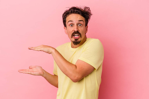 Young caucasian man isolated on pink background shocked and amazed holding a copy space between hands. - Foto, Bild
