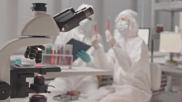 Średni długi męski Afroamerykanin i kobieta kaukascy naukowcy w kombinezonach, okularach i maskach, siedzący w laboratorium, analizujący chemikalia w probówkach - Materiał filmowy, wideo