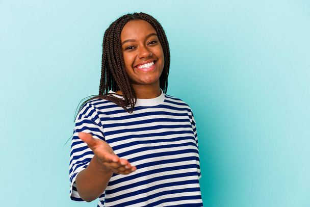 Mladý africký Američan žena izolovaný na modrém pozadí strečink ruku na kameru v pozdrav gesto. - Fotografie, Obrázek