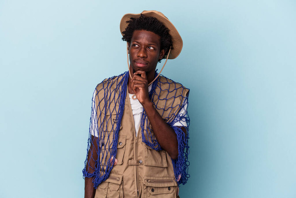 Young african american fisherman holding a net isolated on blue background looking sideways with doubtful and skeptical expression. - Photo, Image