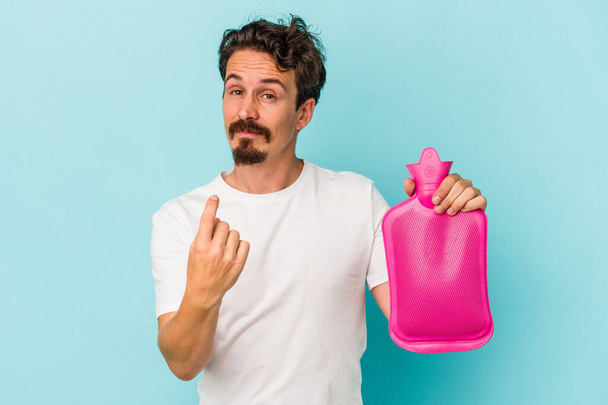 Young caucasian man holding a water bag isolated on blue background pointing with finger at you as if inviting come closer. - Foto, immagini