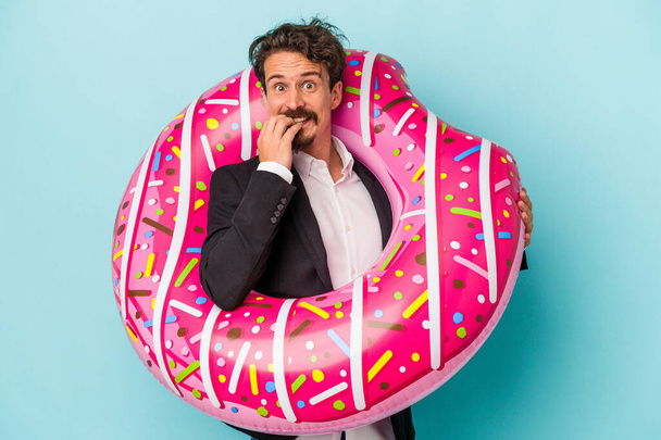 Young business man with inflatable donut isolated on blue background biting fingernails, nervous and very anxious. - Photo, Image