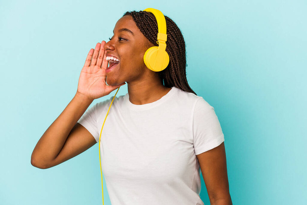 Jonge Afro-Amerikaanse vrouw luisteren naar muziek geïsoleerd op blauwe achtergrond schreeuwen en houden palm in de buurt van geopende mond. - Foto, afbeelding