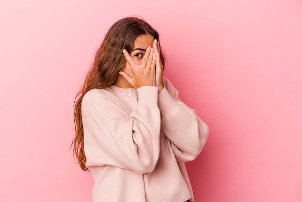Молодая белая женщина, изолированная на розовом фоне, моргает сквозь пальцы испуганными и нервными. - Фото, изображение