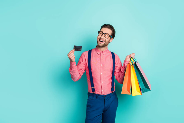 Photo of attractive dreamy guy dressed pink shirt spectacles holding bags card empty space isolated turquoise color background - 写真・画像